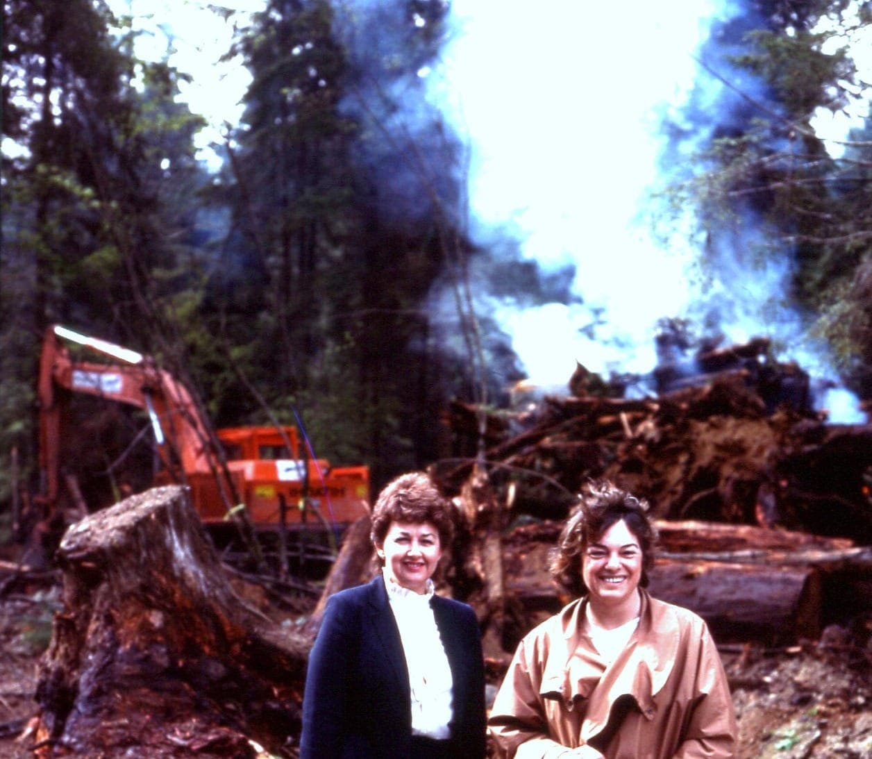Two original members standing at the freshly cleared site, land-clearing fire burning behind them